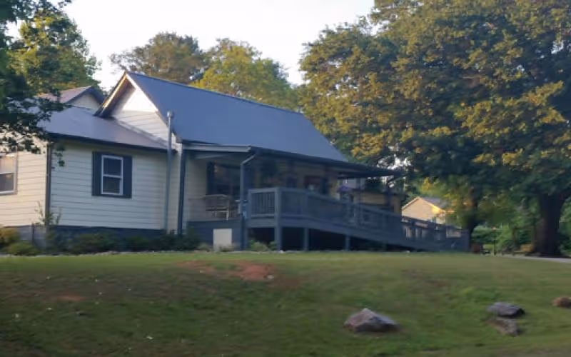 A single-story house with a gray roof and light-colored siding, featuring a front porch with a wheelchair ramp. The house is surrounded by green grass, trees, and some rocks on the lawn.