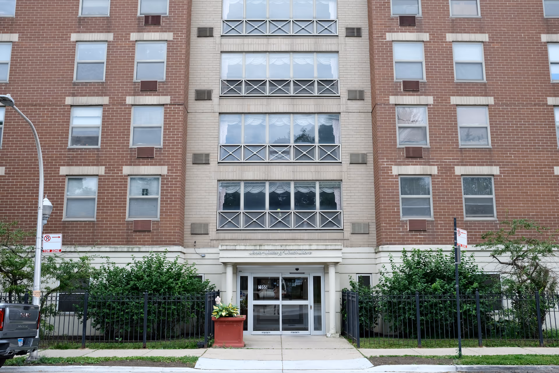 Front facade and entrance of a multi-story brick senior living building with a small fenced garden and potted plants.