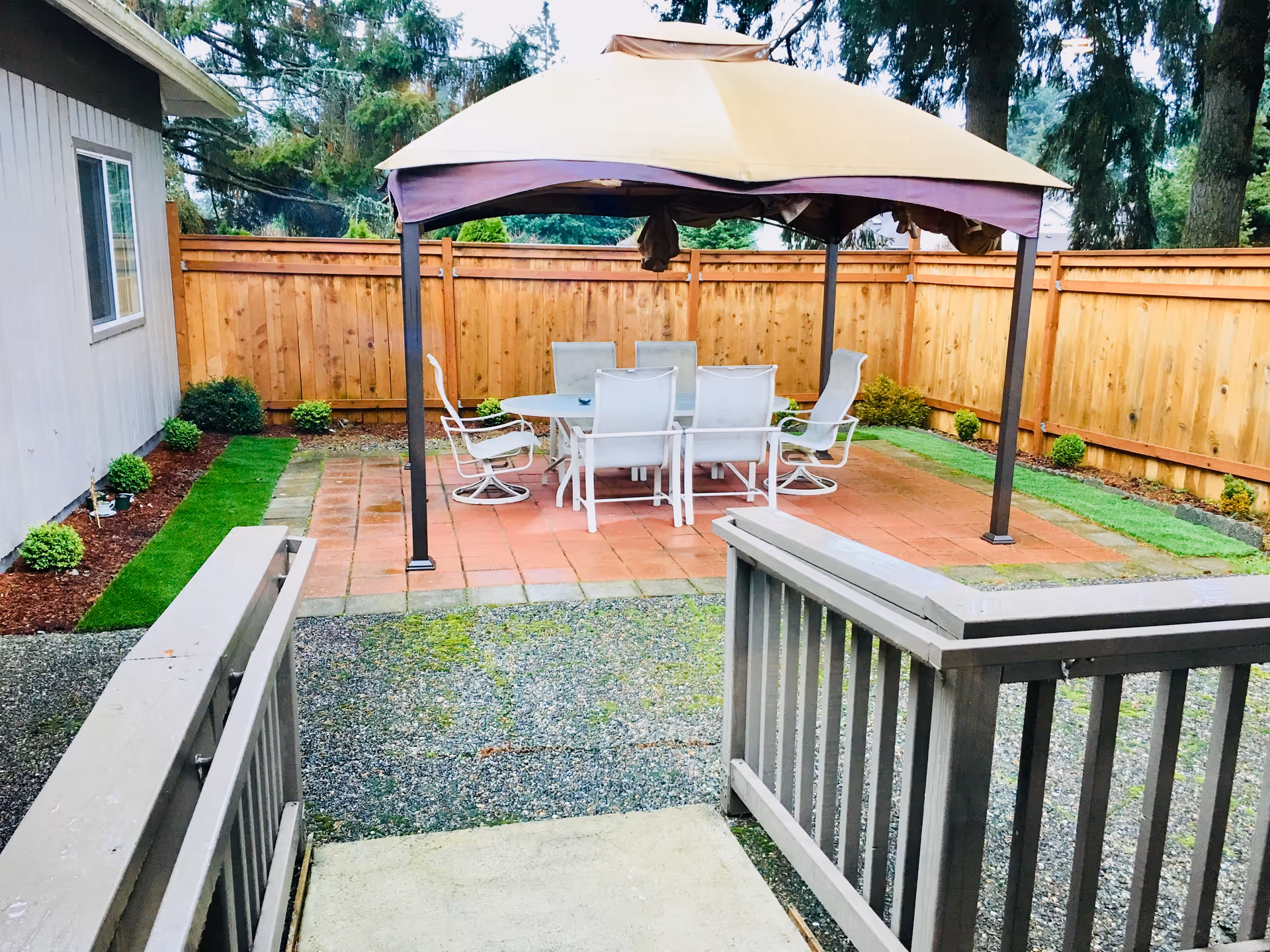 Outdoor patio with a beige gazebo, white table and chairs, surrounded by a wooden fence.