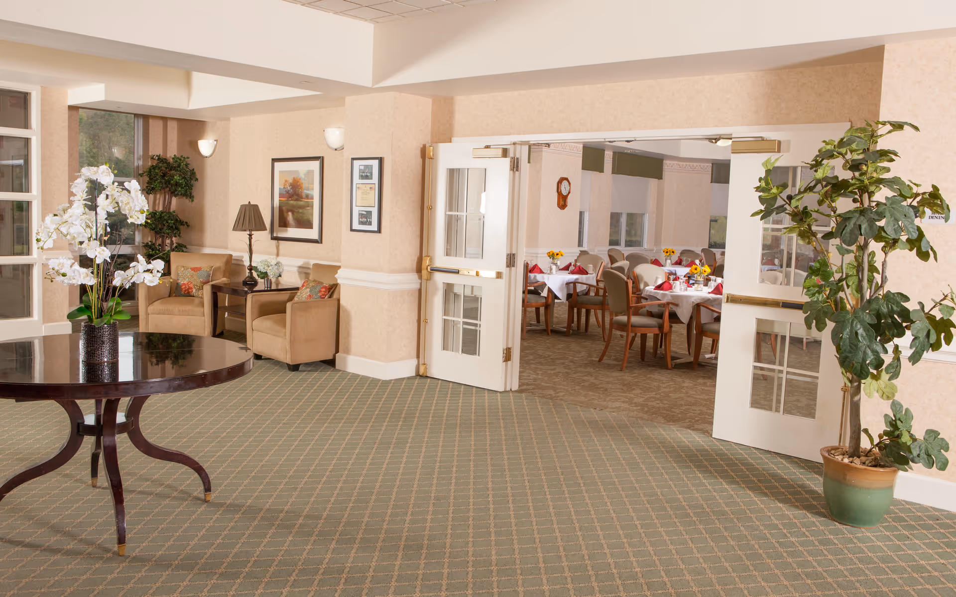 Lobby of a senior living facility with seating, a round table holding flowers, potted plants, and open double doors revealing a dining room.