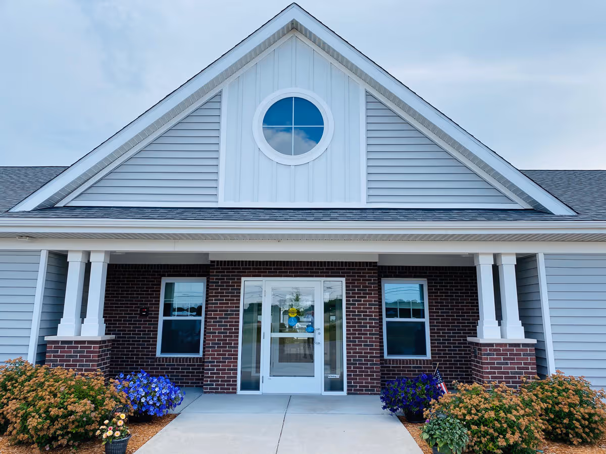 Front exterior view of a senior living facility building with a gabled roof, a circular window above the entrance, brick and siding walls, two windows flanking a glass door, and landscaped bushes and flowers along the walkway.