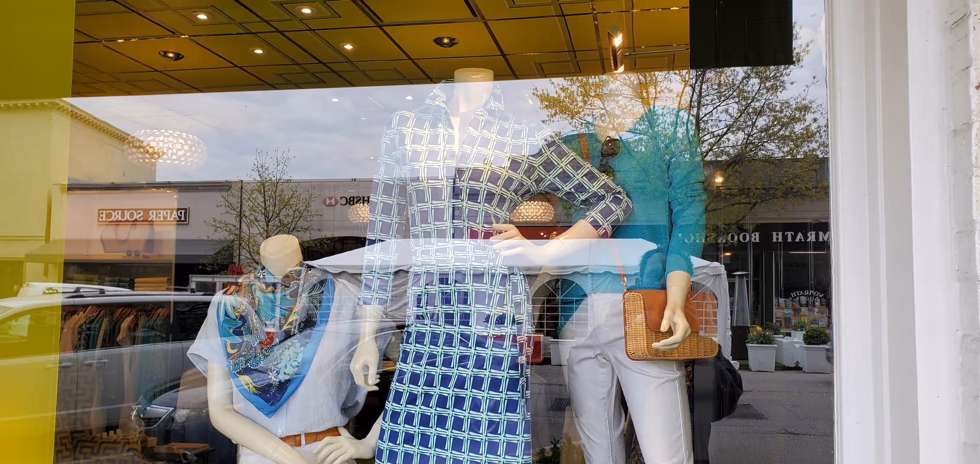 Storefront window display featuring mannequins wearing outfits, with reflections of a street scene.