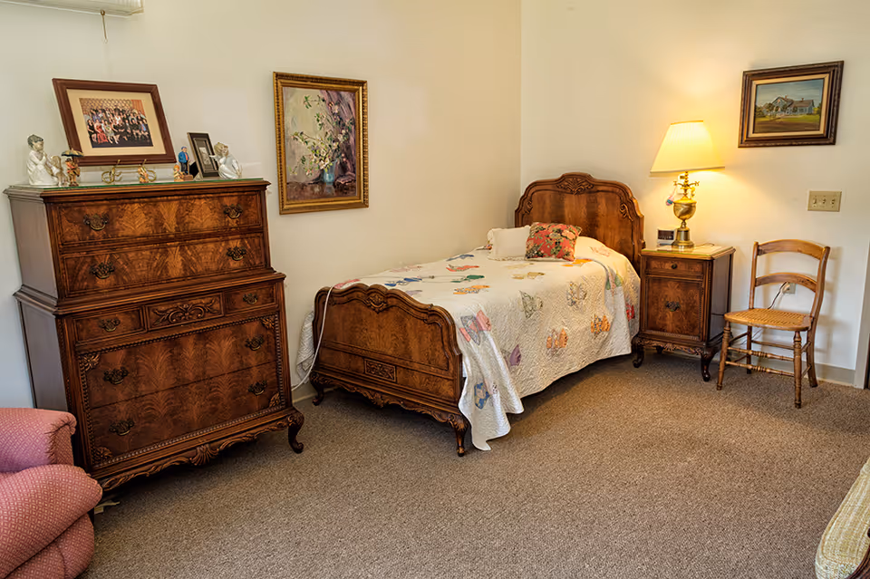 A traditionally furnished bedroom with a single wooden bed, matching dresser and nightstand, a lamp and a chair.