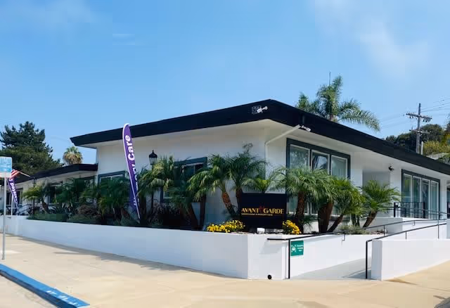 Low-profile white senior living building with palm trees, purple banners, and a sidewalk in front.