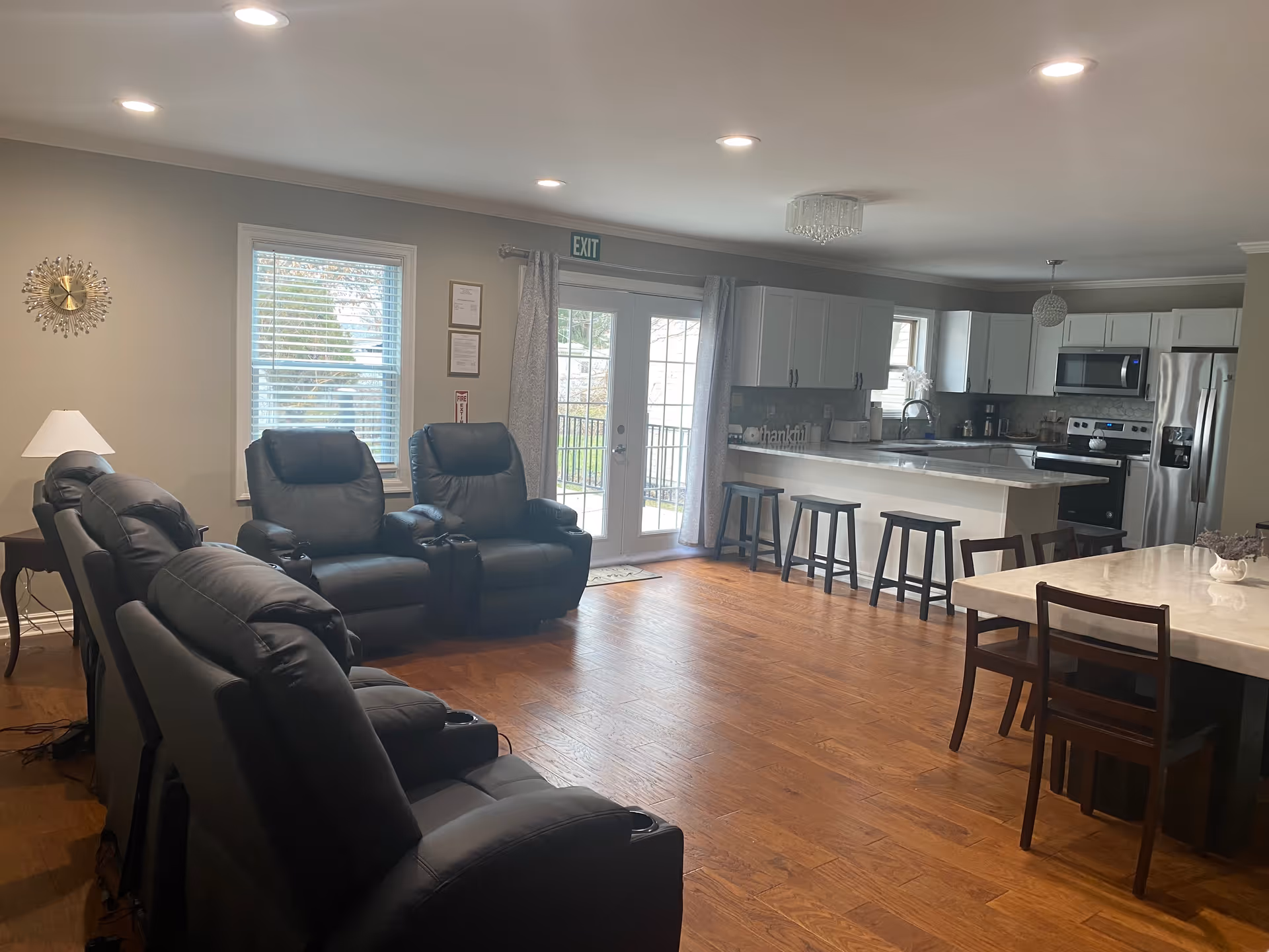 A spacious and well-lit interior room featuring a seating area with multiple black recliner chairs arranged in rows, a window with blinds, a lamp on a side table, and an open kitchen with white cabinets, stainless steel appliances, a kitchen island with four stools, and a dining table with chairs. The room has wooden flooring and neutral-colored walls.