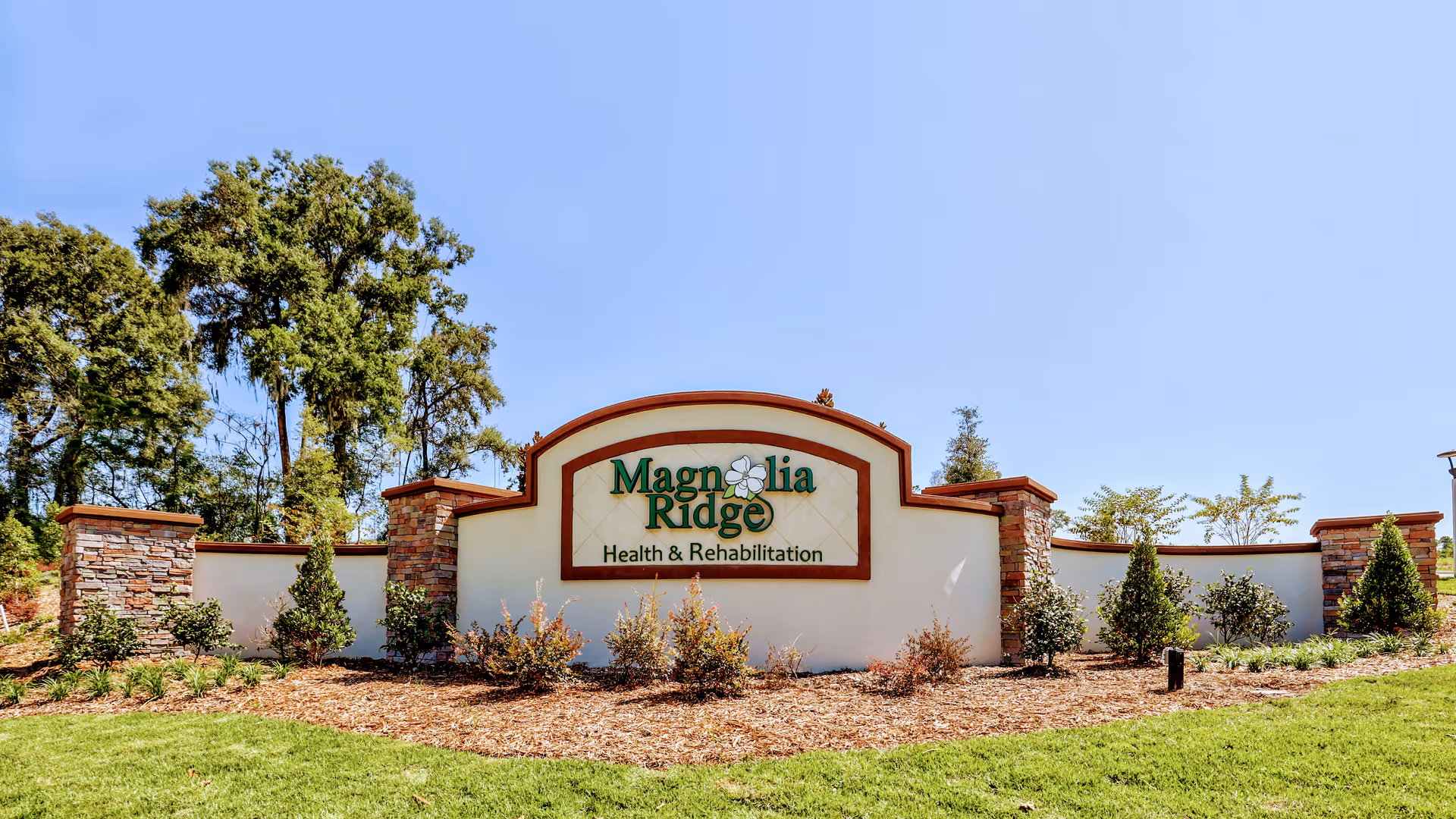 Exterior entrance sign reads Magnolia Ridge Health and Rehabilitation, surrounded by shrubs and trees.