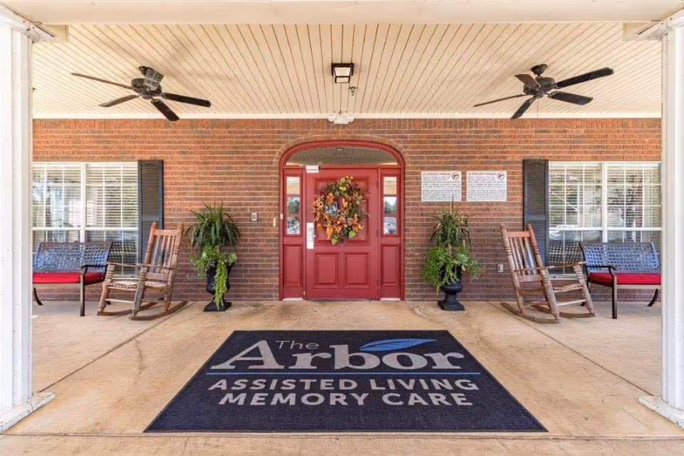 Entrance area of The Arbor Assisted Living and Memory Care facility featuring a covered porch with two ceiling fans, a red front door decorated with a fall-themed wreath, two potted plants on either side of the door, wooden rocking chairs, and benches with red cushions. A large black mat with the facility's name is placed on the ground in front of the door.