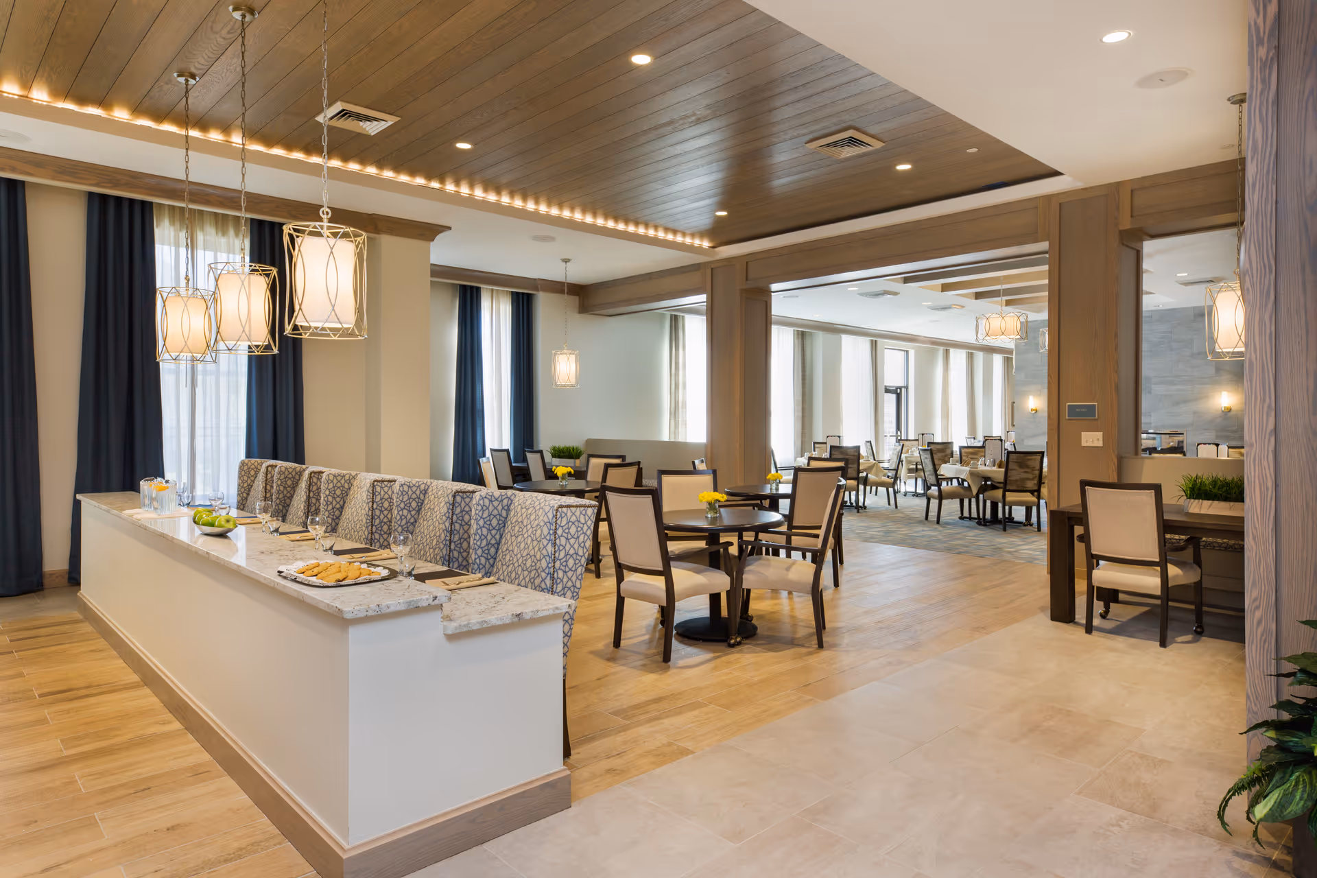 A modern senior living dining area with a long pastry counter, tables, chairs, and warm lighting.