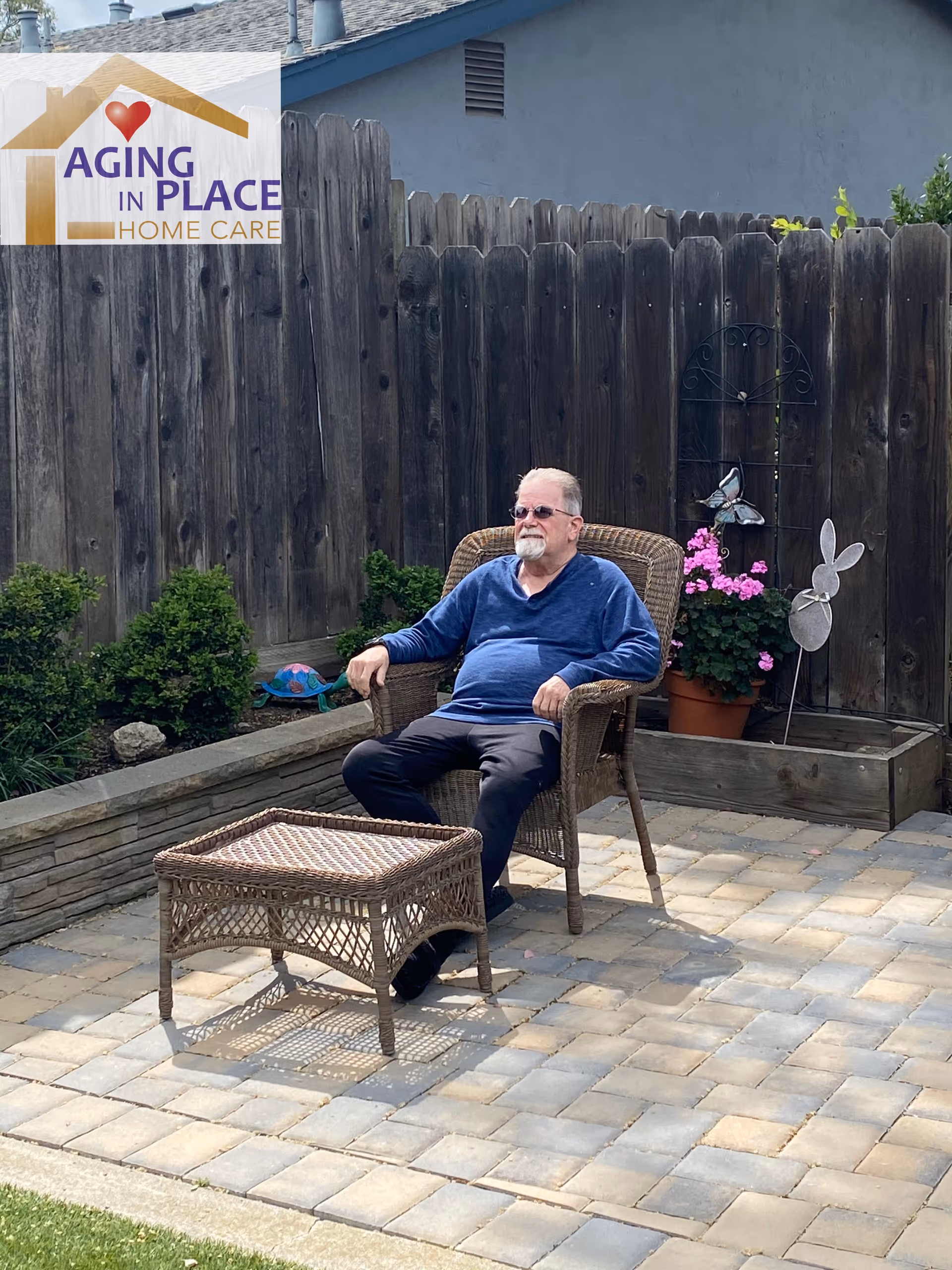 Elderly man sits in wicker chair beside Aging in Place Home Care sign on patio.