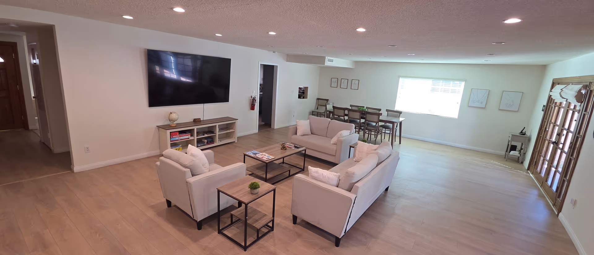 Spacious communal living room with beige sofas, coffee tables, and a wall-mounted TV.