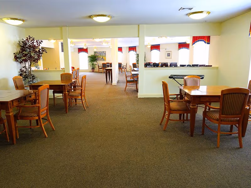 A spacious, well-lit common area with multiple wooden tables and chairs arranged for seating. The room has beige walls, carpeted floors, ceiling lights, and decorative plants. A keyboard is placed on a ledge dividing the room, and windows with red and blue valances allow natural light to enter.