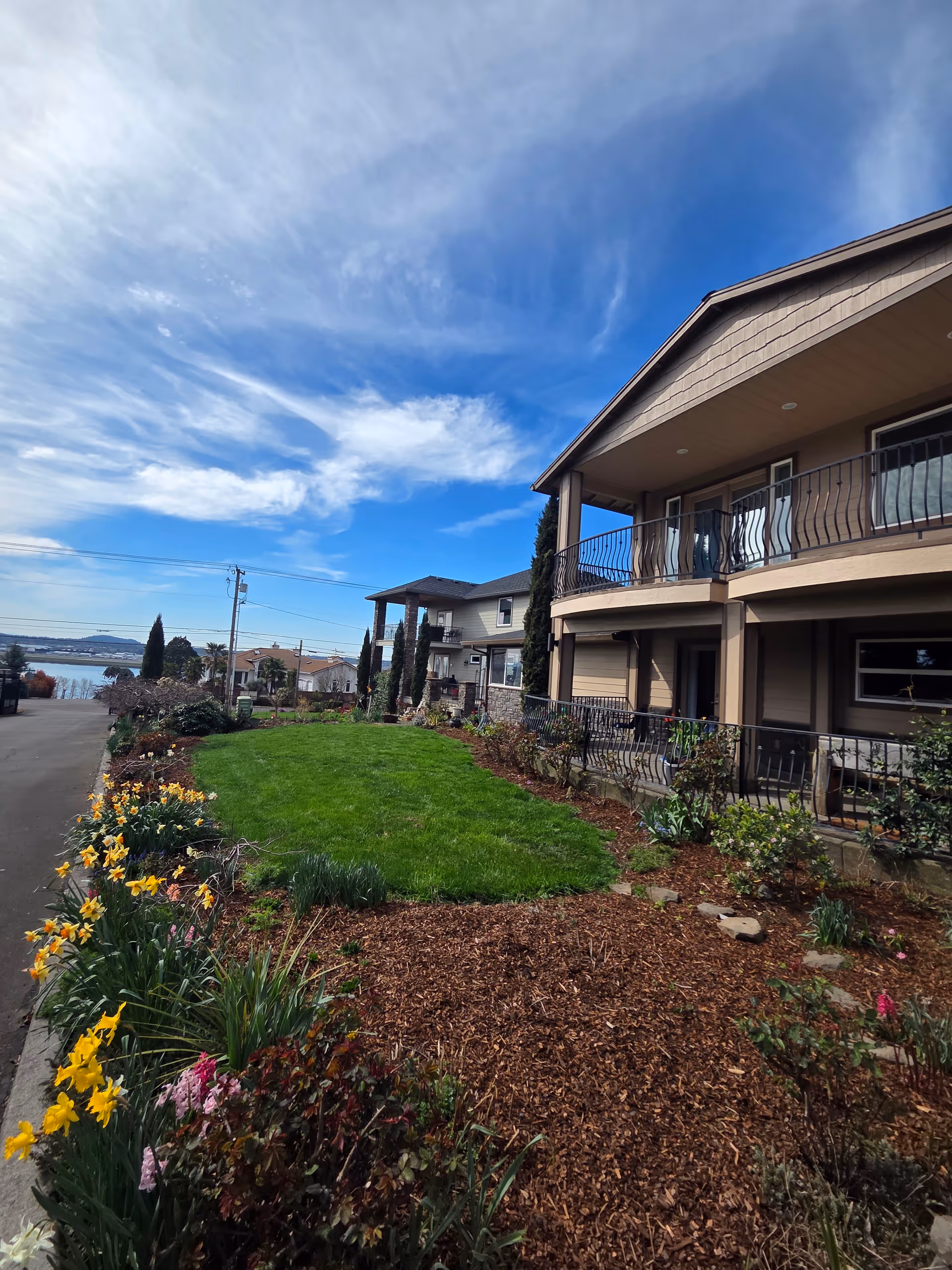 Beige multi-story senior home with balconies, a manicured lawn, and colorful flower beds along the walkway.