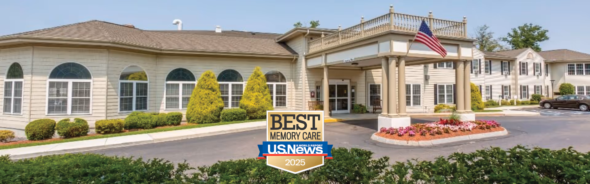 Exterior view of Benchmark Senior Living at Waltham Crossings building with a covered entrance, landscaped bushes, an American flag, and a badge indicating Best Memory Care by U.S. News 2025.