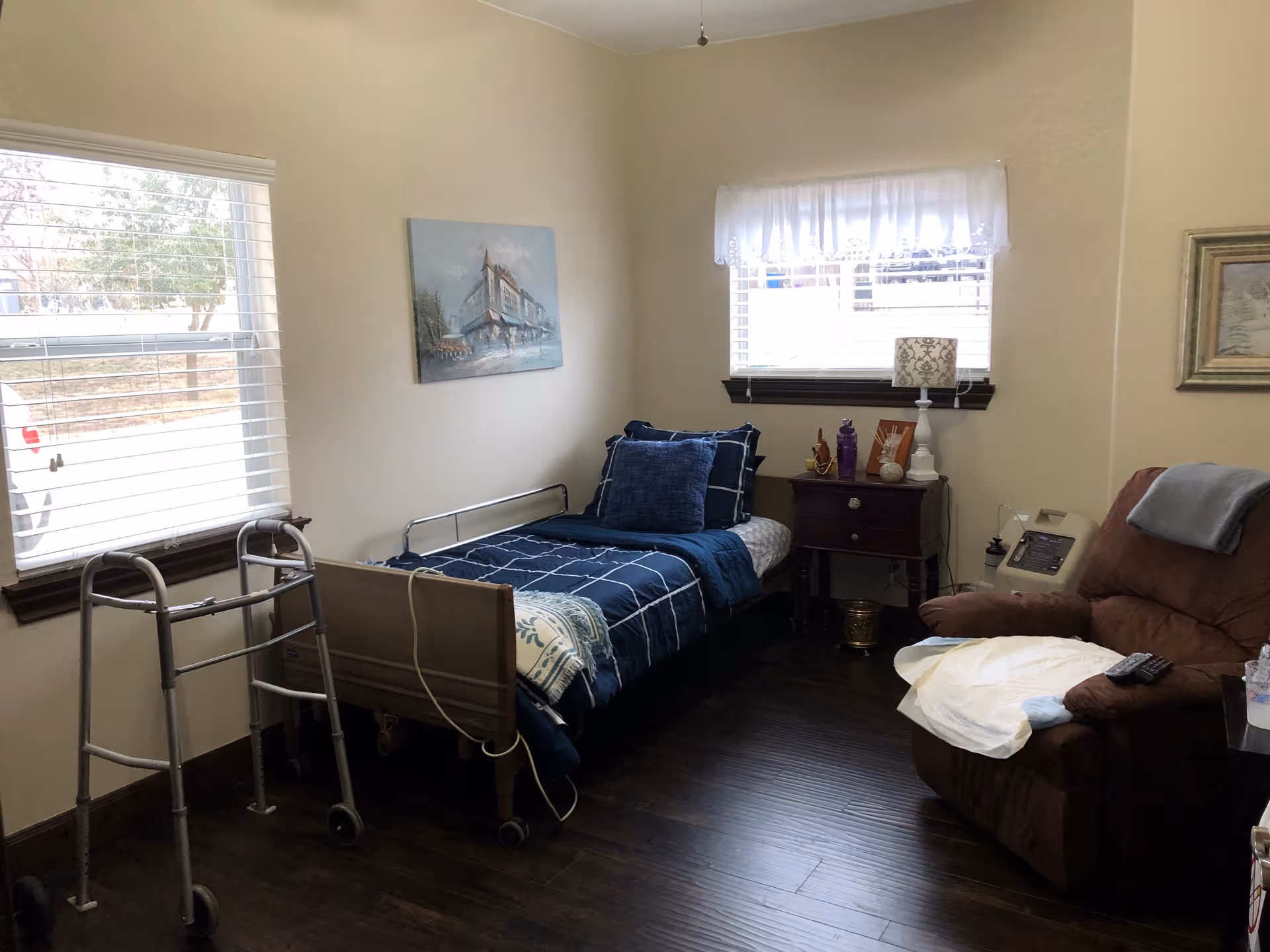 A small, cozy bedroom in an assisted living facility with a single bed covered in blue bedding, a walker positioned near the window, a wooden nightstand with a lamp and decorative items, and a comfortable brown recliner chair with a remote control on the armrest. The room has two windows with blinds and light-colored walls adorned with framed artwork.