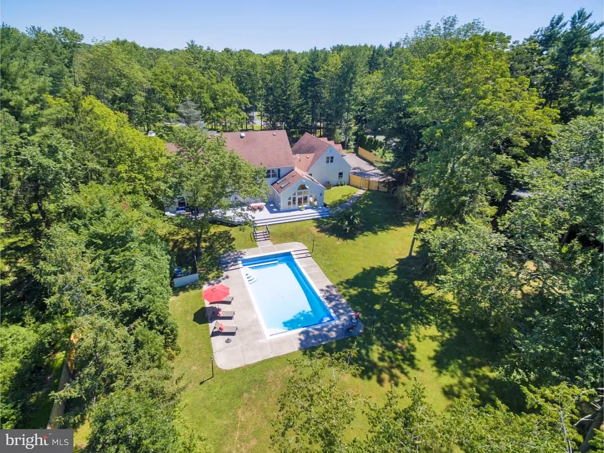Aerial view of a residential property surrounded by dense green trees. The property features a large house with a red roof, a spacious backyard with a rectangular swimming pool, several lounge chairs with red cushions, and a red umbrella providing shade. The area is well-maintained with a concrete pool deck and a grassy lawn.
