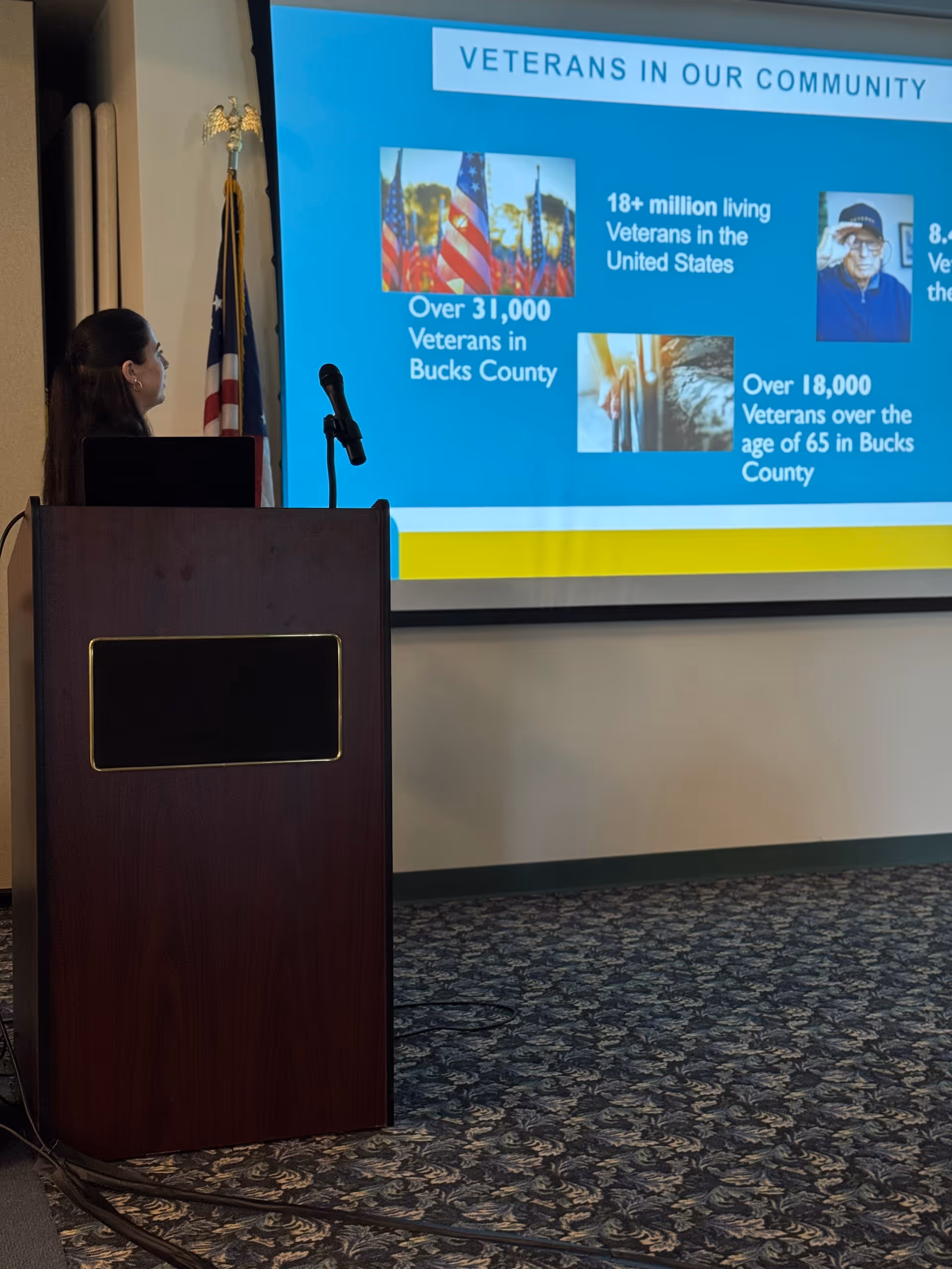Speaker stands at a wooden podium presenting beside a large screen displaying veterans infographic indoors.