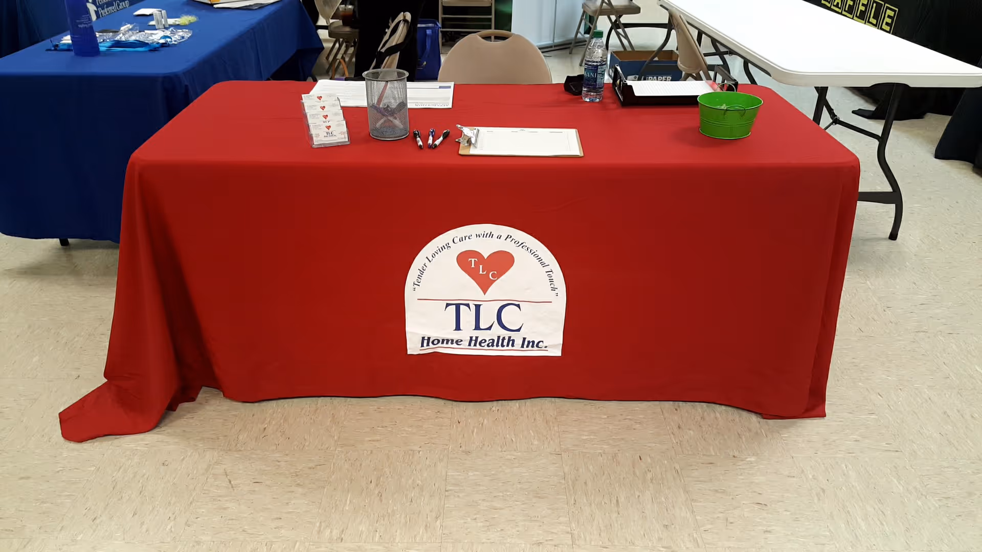 A red tablecloth-covered display table with TLC Home Health Inc. logo at an indoor event.