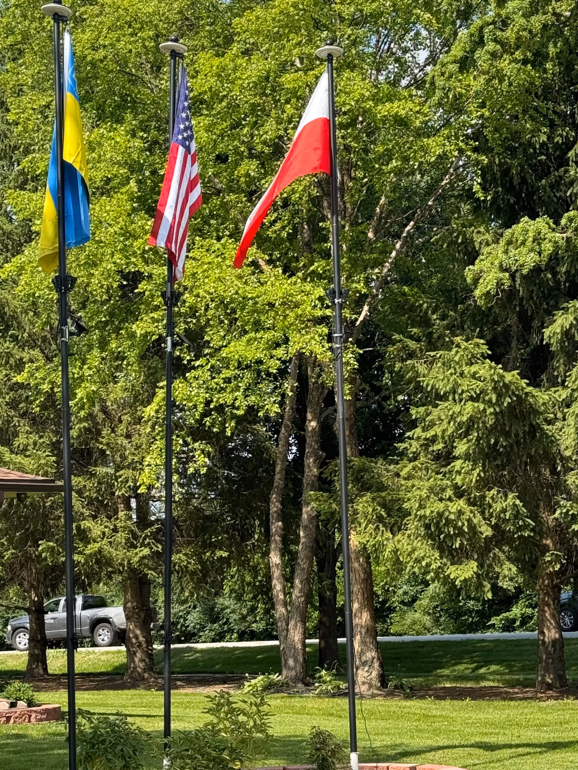 Flagpoles with Ukrainian, American, and Polish flags in a grassy, tree-lined outdoor area.