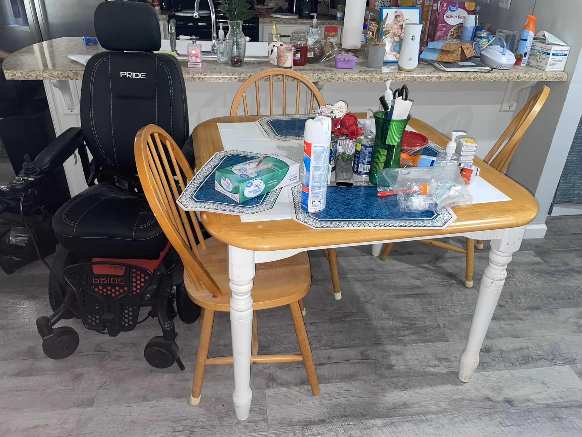 A cluttered dining table with chairs and a wheelchair in a bright kitchen area.