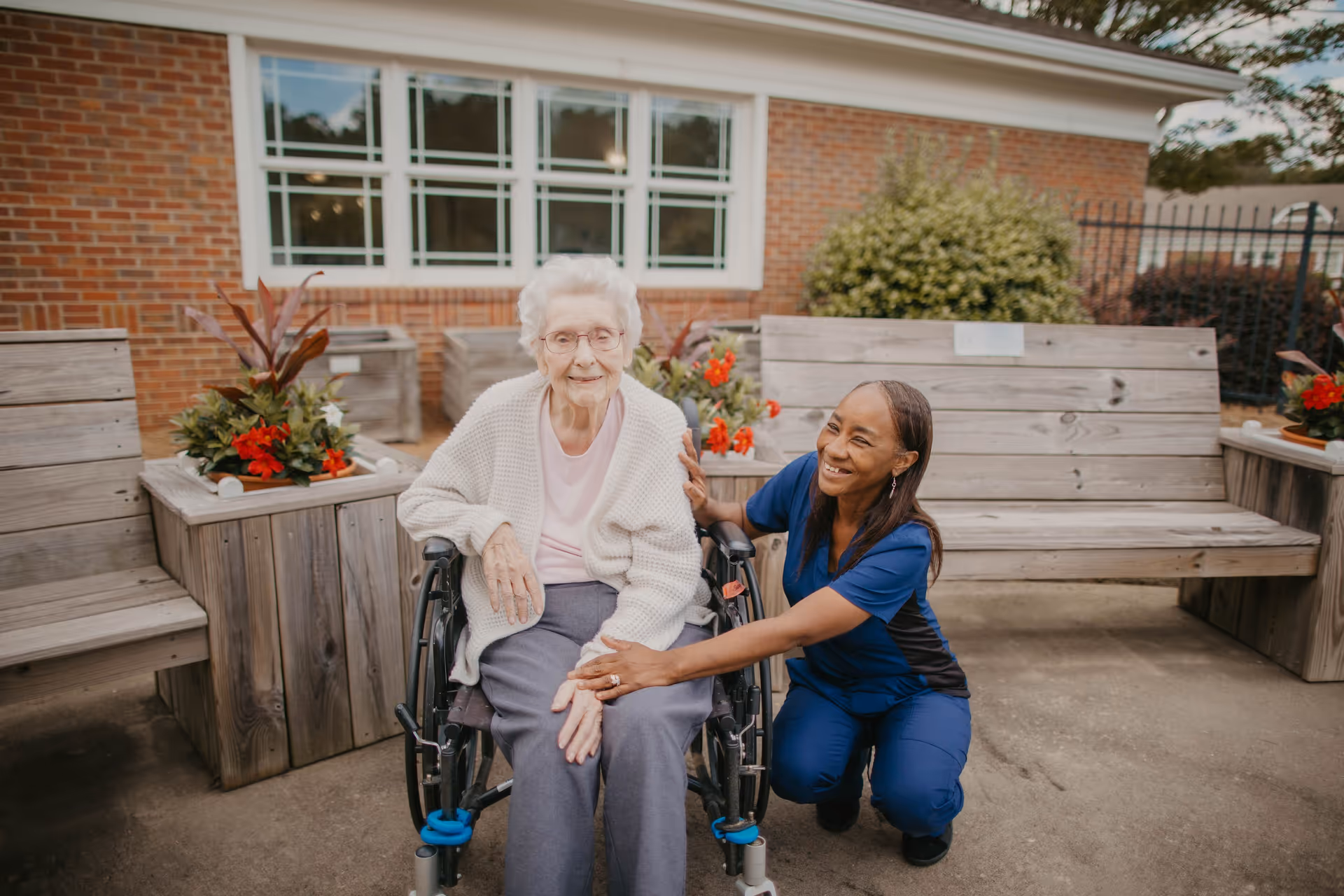 An elderly woman in a wheelchair receives assistance from a caregiver outdoors near planters.