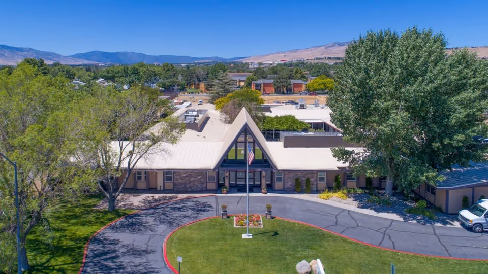 Aerial view of Rosewood Rehabilitation Center showing a single-story building with a distinctive triangular roof entrance, surrounded by trees and greenery. There is a circular driveway with a flagpole and landscaped flower bed in the center. Mountains and other buildings are visible in the background under a clear blue sky.