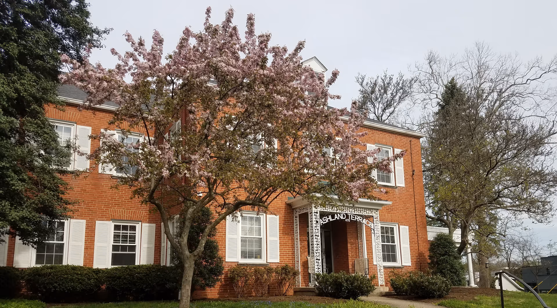 Exterior view of a two-story red brick building with white window shutters and a white decorative entrance canopy labeled 'Ashland Terrace'. In front of the building, there is a tree with pink blossoms and some green shrubs.