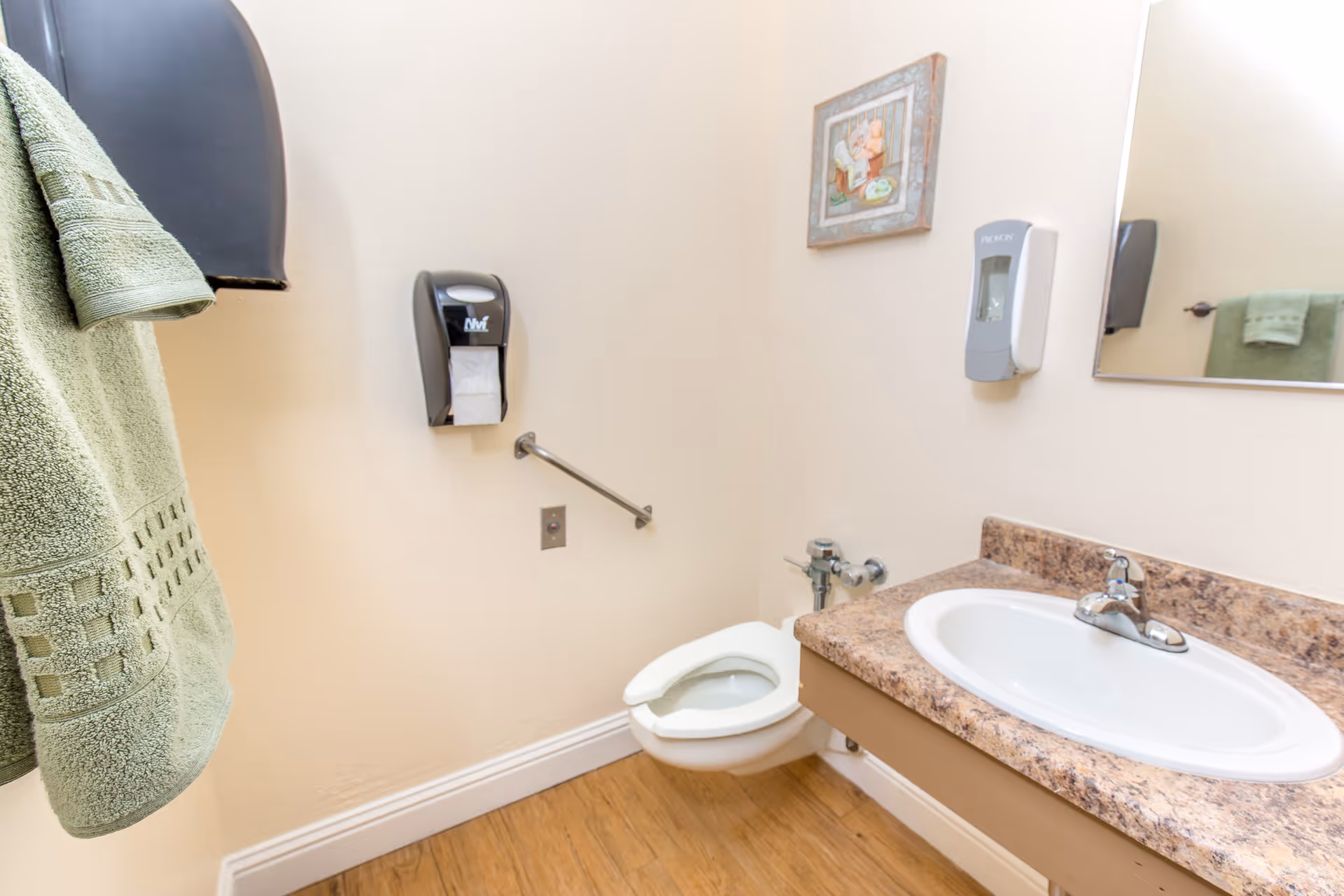 A small bathroom with a wall-mounted toilet, a sink with a marble-patterned countertop, a mirror above the sink, a soap dispenser on the wall, a paper towel dispenser, a green towel hanging on a rack, and a small framed picture on the wall.