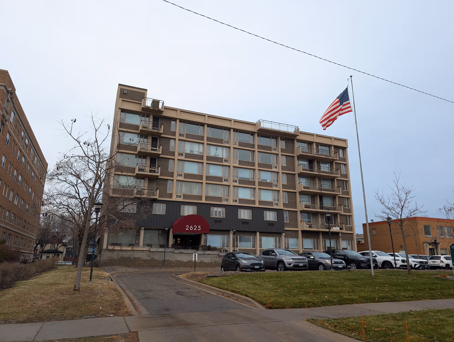 Exterior view of a multi-story senior living facility building with a maroon awning displaying the number 2625. Several cars are parked in front of the building, and an American flag is flying on a flagpole to the right. Leafless trees and a grassy area surround the driveway leading to the entrance.