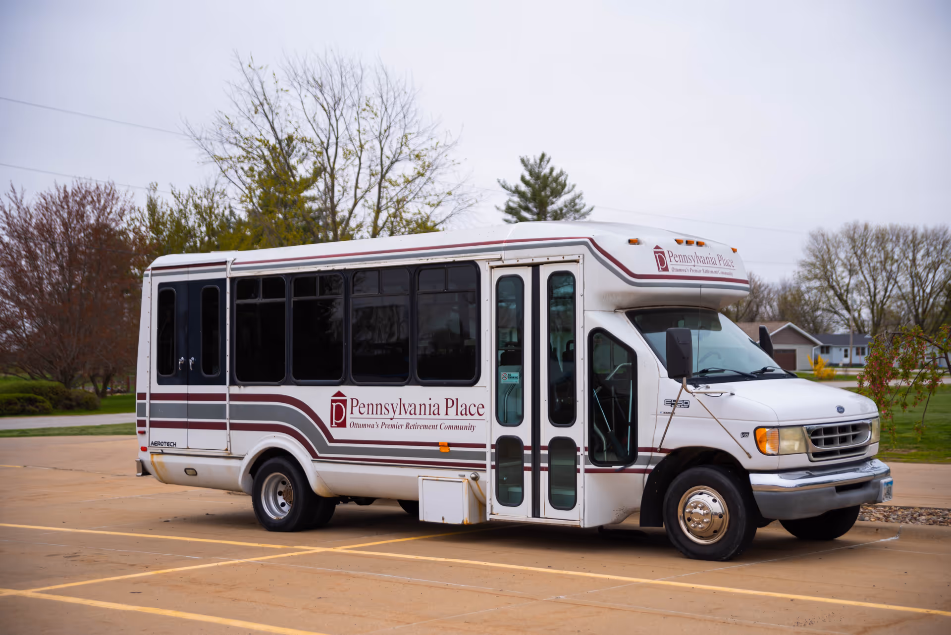 White Pennsylvania Place bus parked in a lot with trees and suburban houses behind.