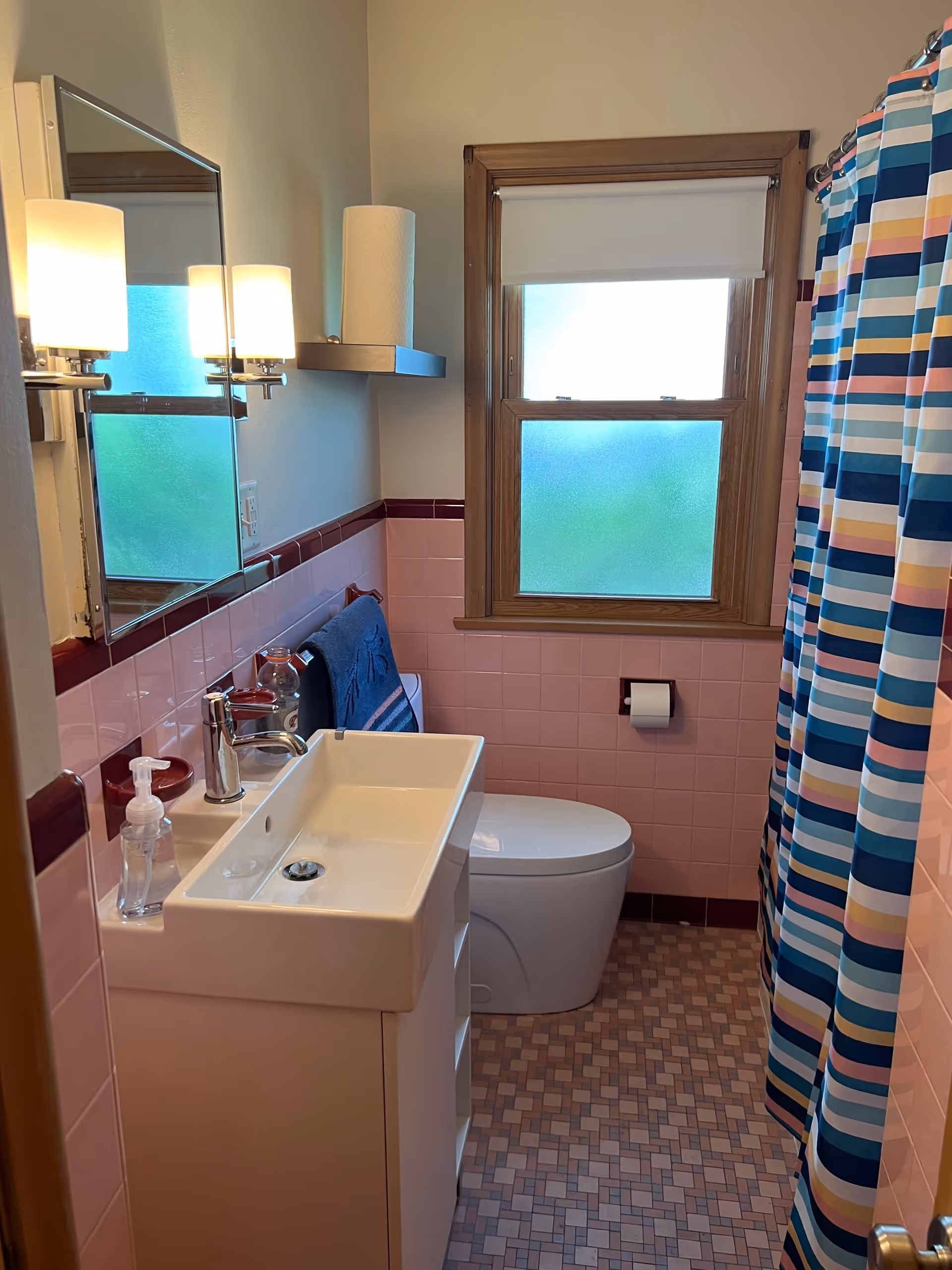 Compact bathroom with pink tile walls, white sink, toilet, striped shower curtain, frosted window.