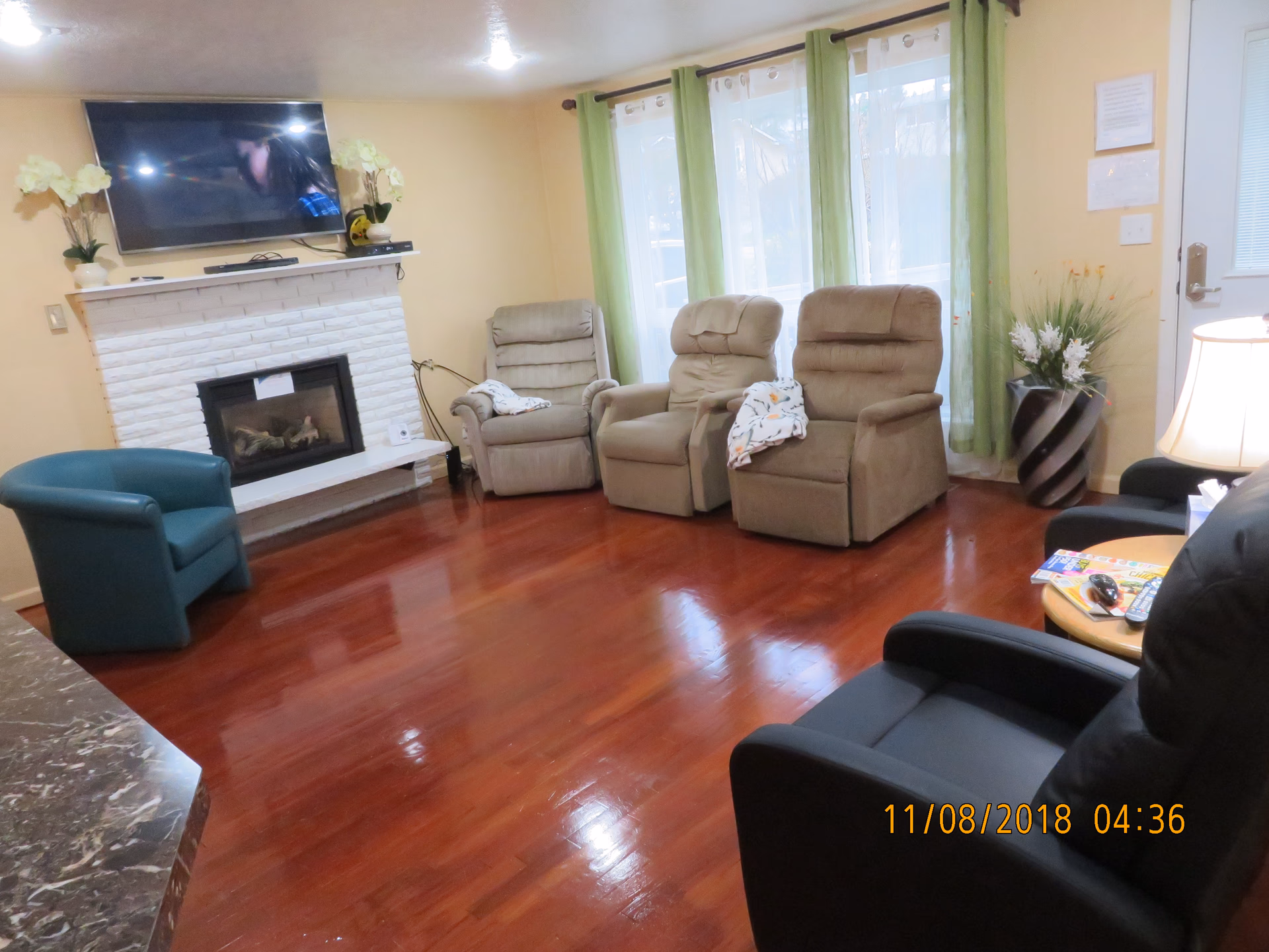 A cozy living room with polished wooden floors, a white brick fireplace with a mounted flat-screen TV above it, three beige recliner chairs with patterned blankets, a teal armchair, a black armchair, a small round wooden table with magazines and remote controls, green curtains on large windows, and a decorative vase with flowers near a door.