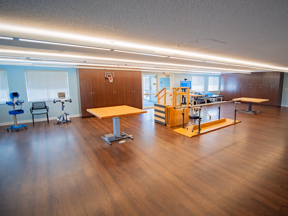 Spacious interior rehab room with parallel bars, therapy tables, and a wooden floor.