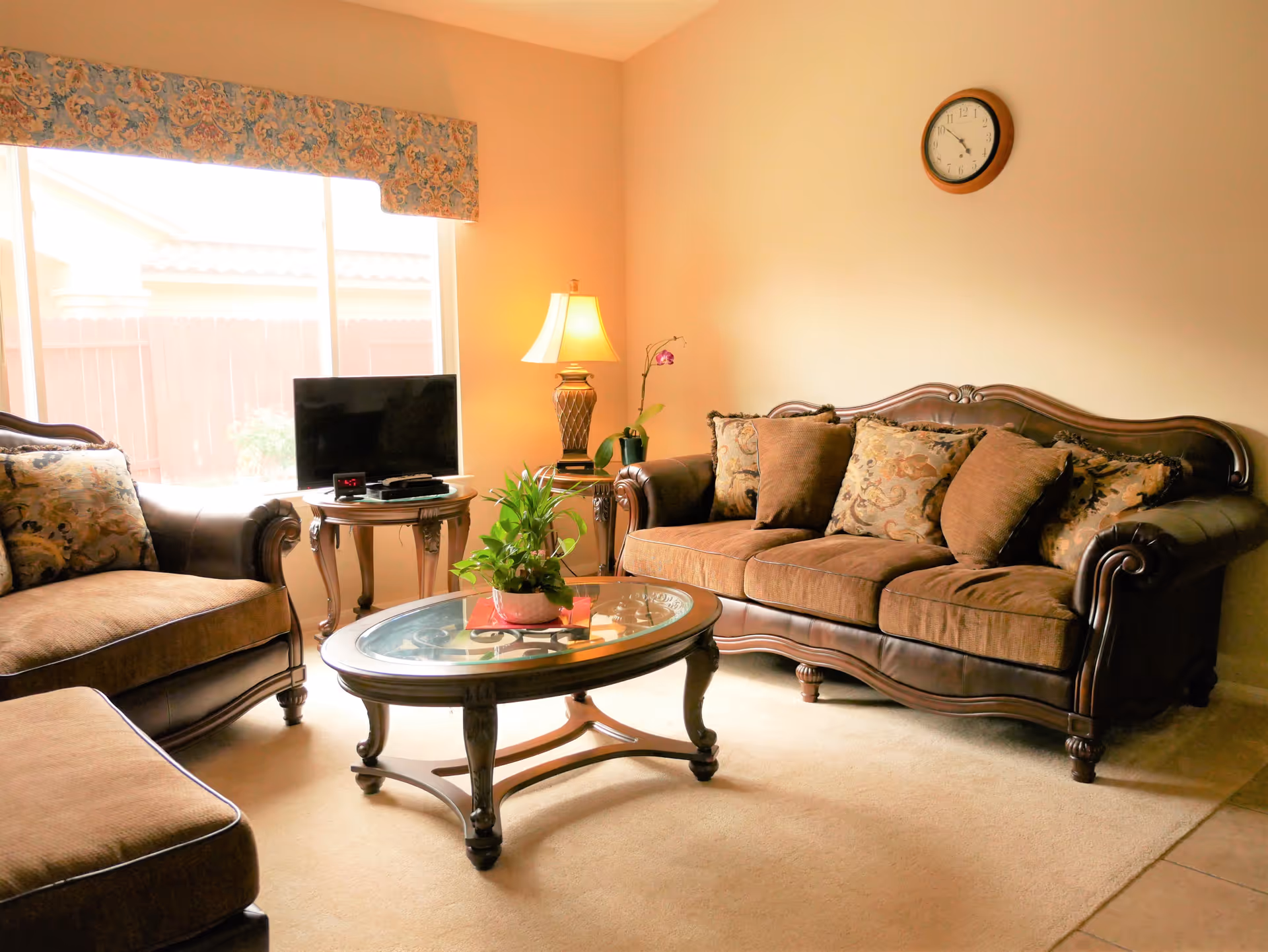 A cozy living room with a brown upholstered sofa and matching armchair, a glass-top wooden coffee table with a potted plant, a side table with a lamp and a small TV, and a large window with floral valance letting in natural light. A round wall clock is mounted on the beige wall.