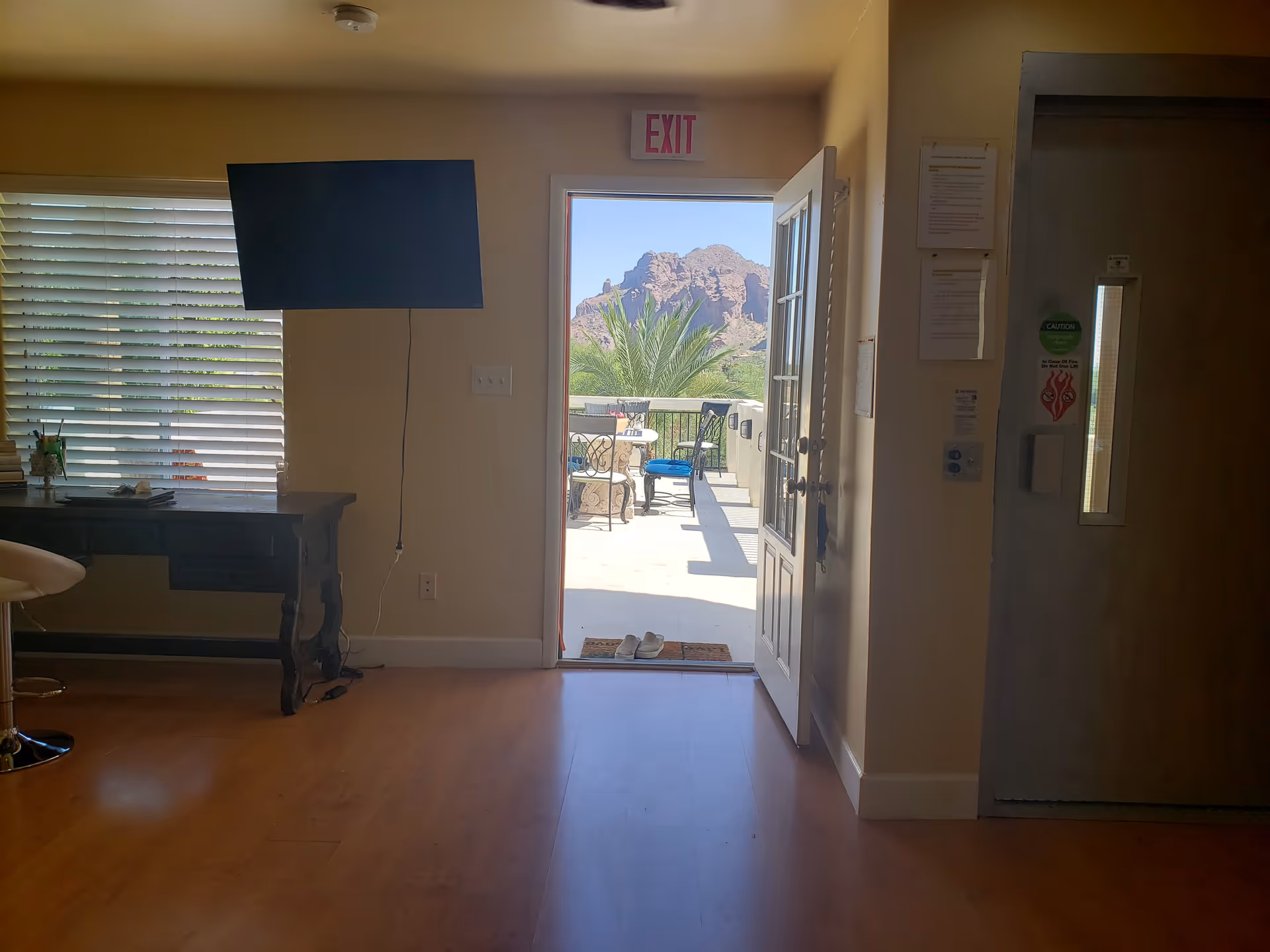 Sunlit living area with TV, desk, and door opening to outdoor patio overlooking mountains.