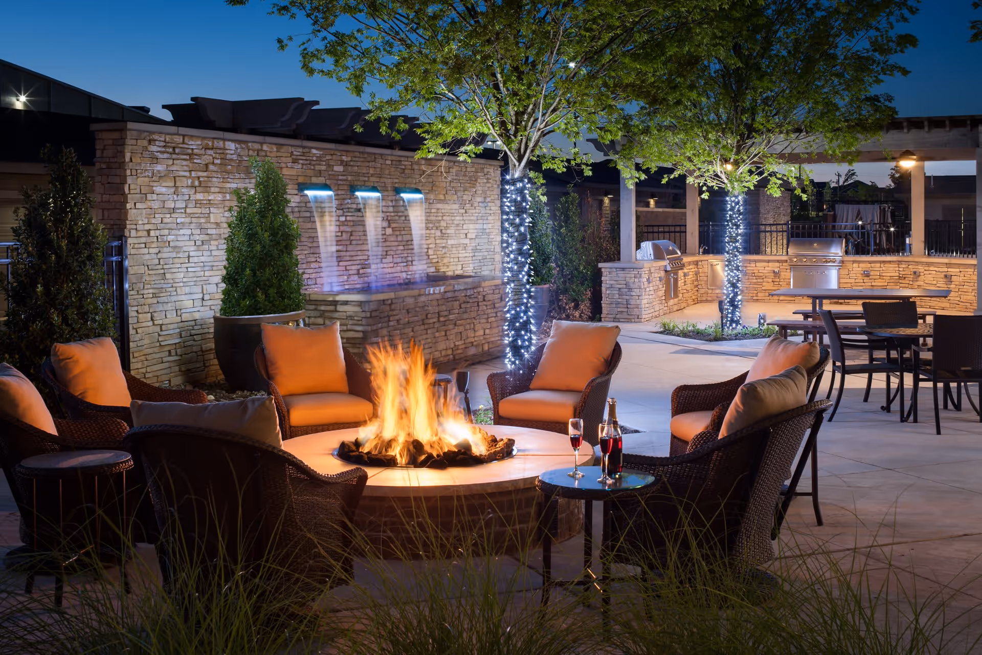 Outdoor patio area at night with a circular fire pit surrounded by cushioned wicker chairs. Trees wrapped with string lights and a stone wall with three water spouts creating a waterfall feature. There are tables and chairs in the background along with built-in grills under a pergola.
