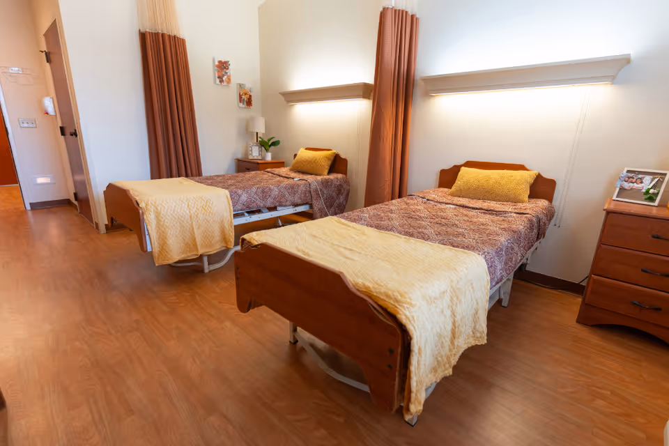 A nursing facility bedroom with two single beds, each with a patterned bedspread and a yellow blanket folded at the foot. There are brown curtains behind each bed, a wooden nightstand with a lamp and a plant between the beds, and a wooden dresser with a framed photo on top. The room has wooden flooring and soft lighting above each bed.