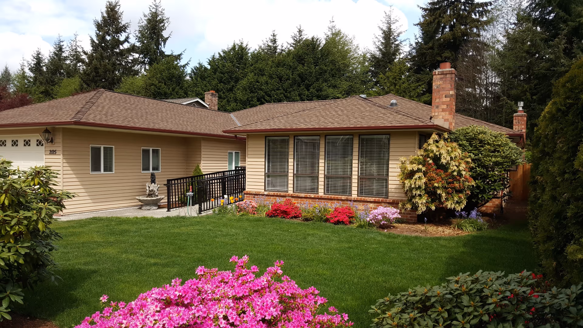 Single-story beige house with large front windows and colorful flowering shrubs in a green yard.