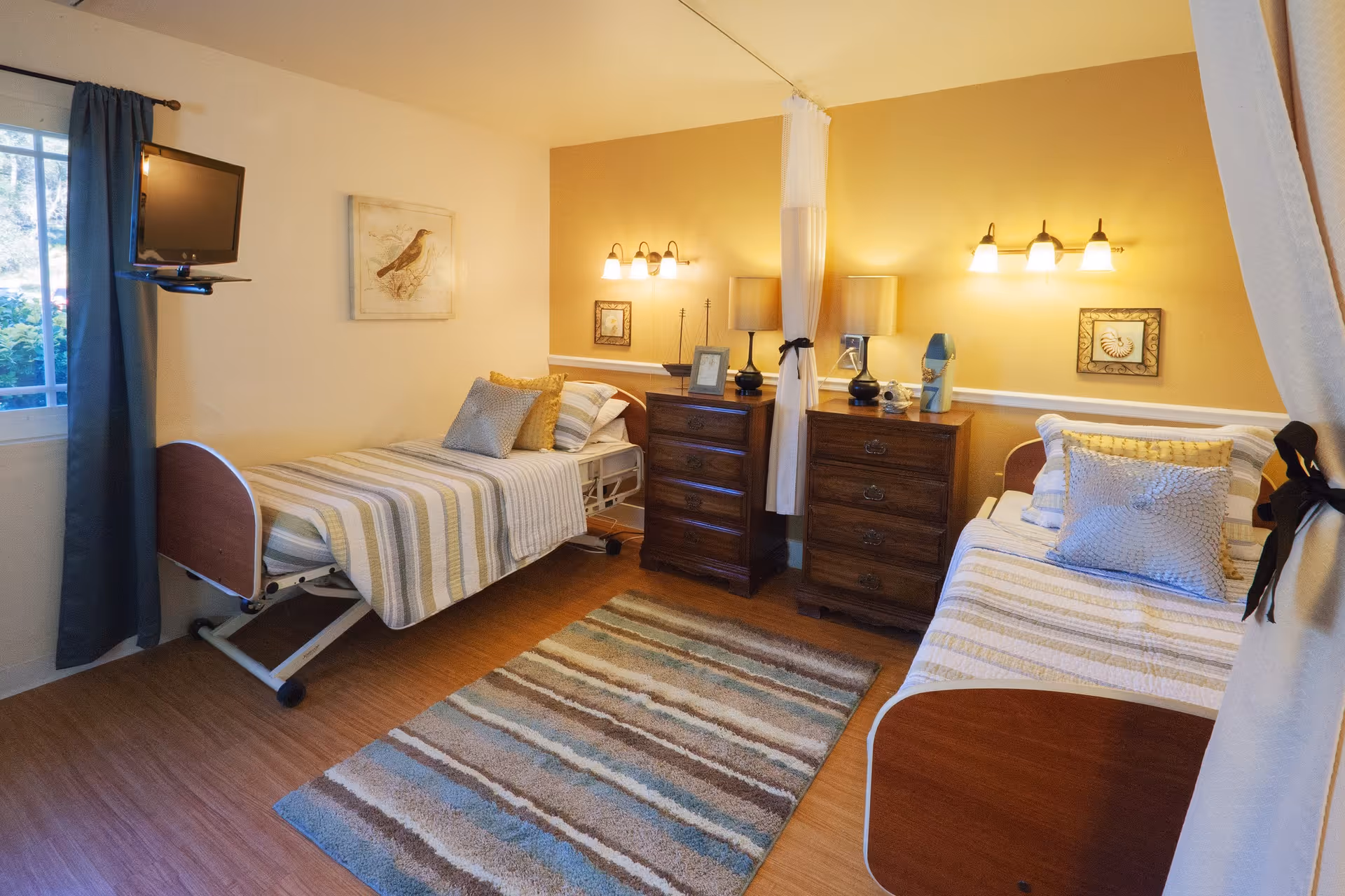 A cozy room with two single beds separated by a curtain. Each bed has striped bedding and multiple pillows. Between the beds are two wooden dressers with lamps and decorative items on top. The walls are painted in warm tones with framed artwork. A window with blue curtains is on the left side, and a flat-screen TV is mounted near the window. A striped area rug covers part of the wooden floor.