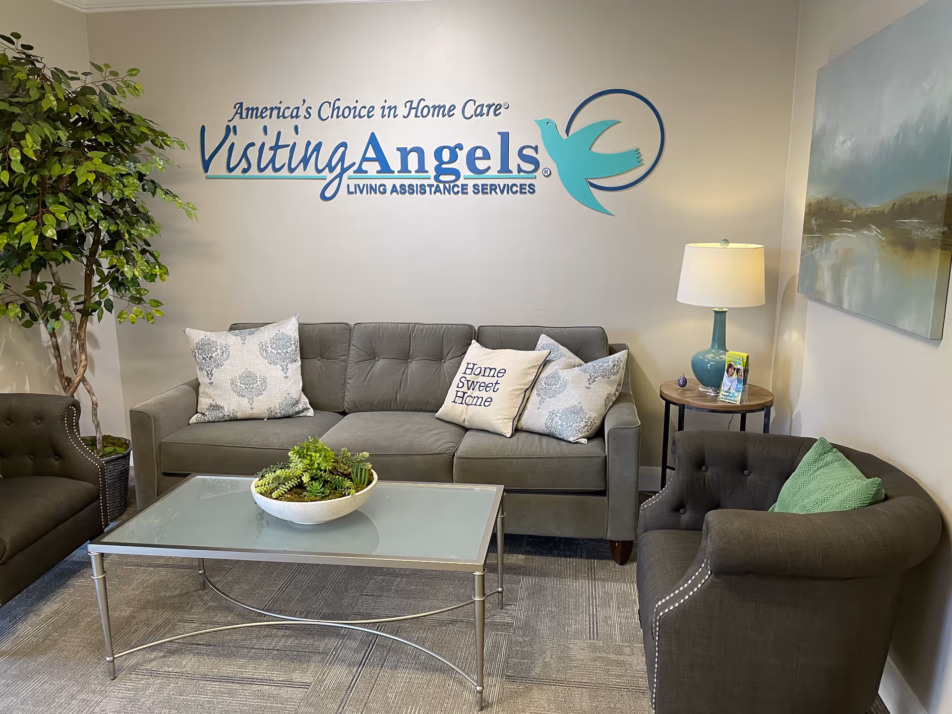 A modern waiting area with gray sofa, glass coffee table, and a Visiting Angels mural.