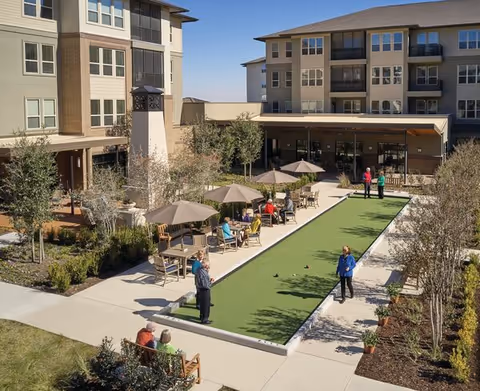 Outdoor courtyard area at Highland Springs Senior Living Community with a bocce ball court in the center. Several seniors are playing or sitting around the court under umbrellas and on benches. The courtyard is surrounded by multi-story residential buildings with large windows and balconies. Trees and landscaped plants line the walkways.