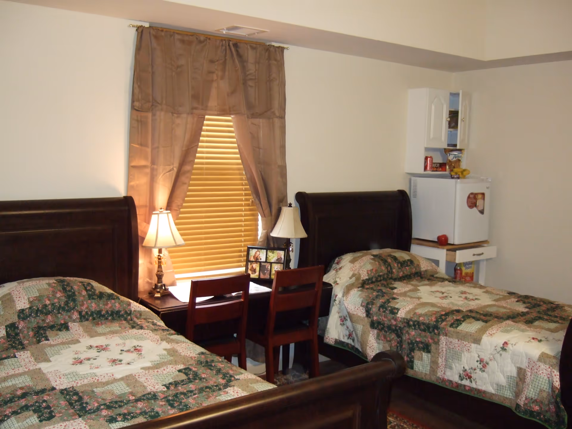 A furnished bedroom with two twin beds with wooden headboards, quilted bedspreads, a window with blinds and curtains, bedside lamps and a small fridge and cabinet.