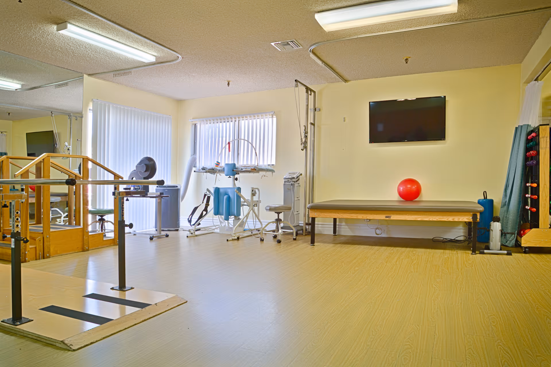 Bright rehabilitation/exercise room with parallel bars, treatment table, exercise equipment and a red therapy ball.
