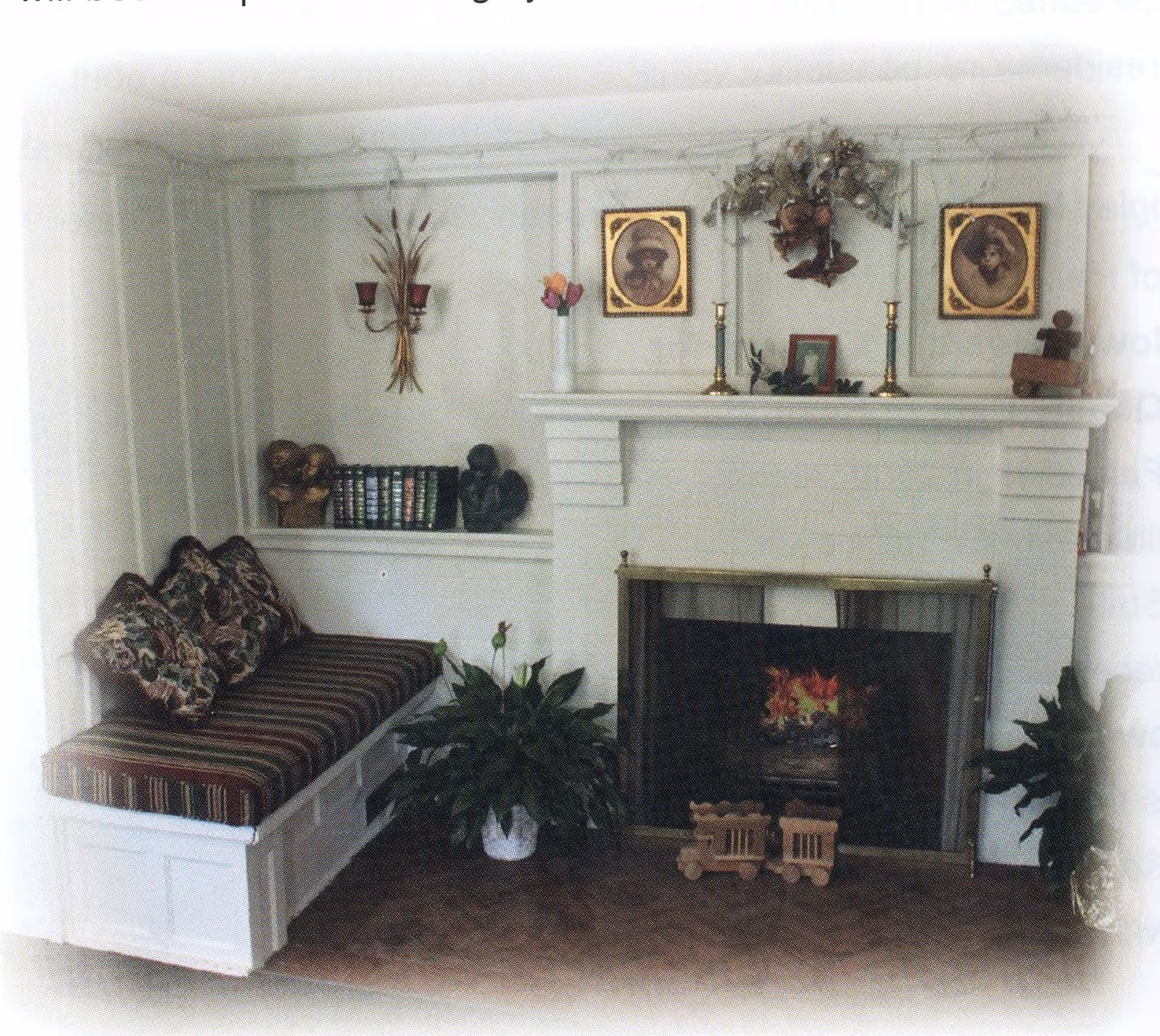 Cozy interior corner featuring a white built-in bench with striped cushions and floral pillows, a white fireplace with a fire burning inside, decorative items on the mantel including framed pictures, candlesticks, and a floral arrangement, potted plants on the floor, and a small wooden toy train in front of the fireplace.