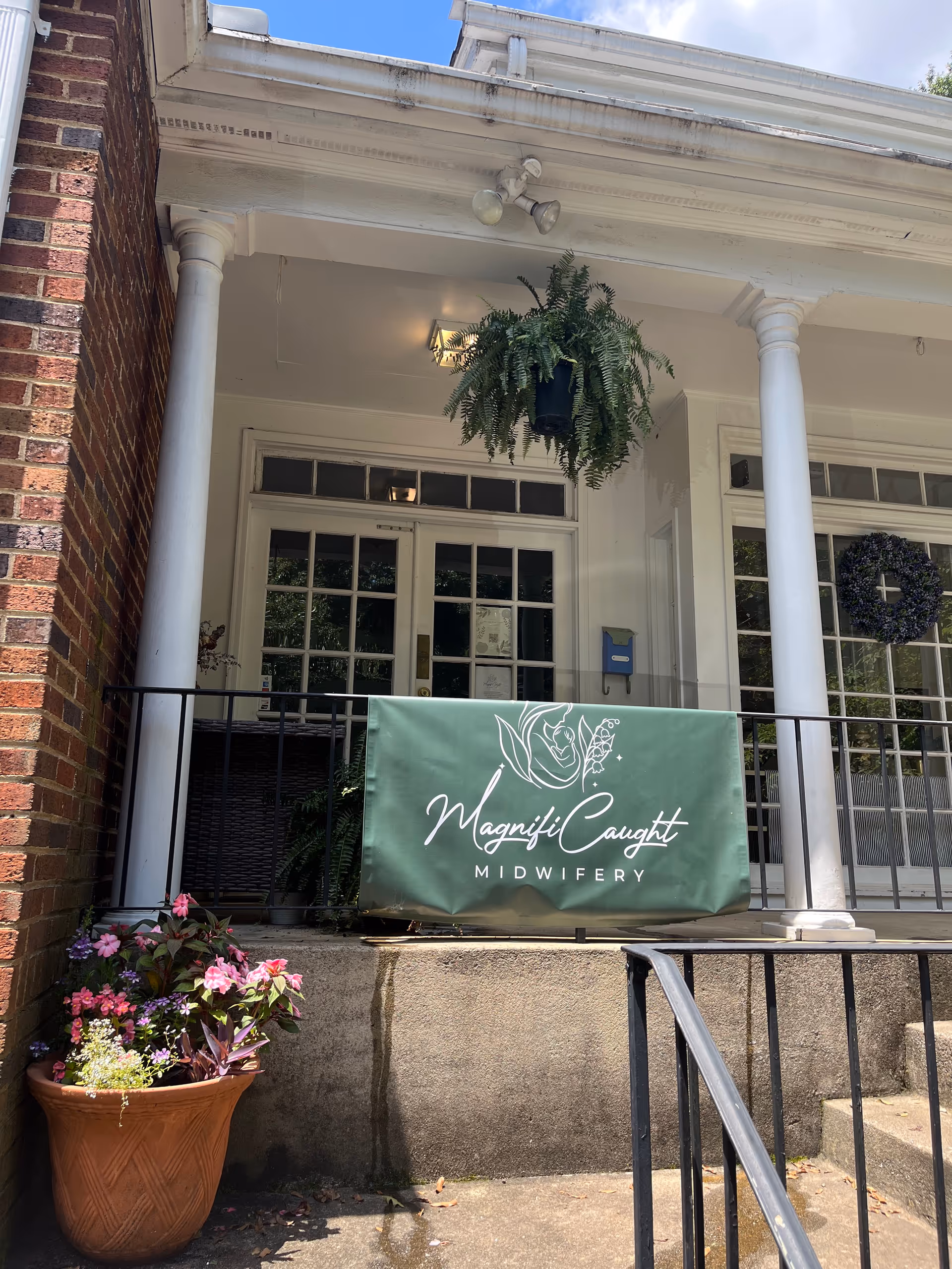 Front entrance of a white-columned building with hanging fern, potted flowers, and MagnifiCaught banner.