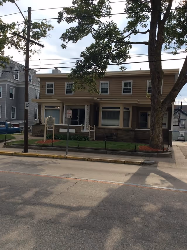 Exterior view of a two-story residential building with beige siding and multiple windows, surrounded by a small lawn with a tree in front. There are utility poles and wires visible, and a street with a sidewalk in the foreground.