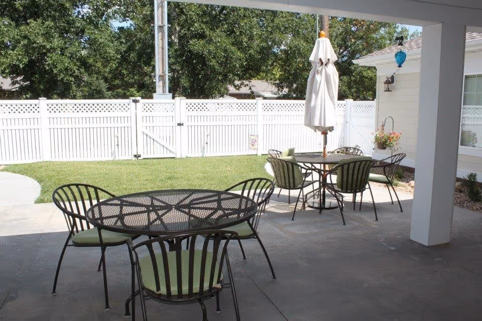 Covered patio with metal tables and chairs and an umbrella overlooking a grassy backyard and white fence.
