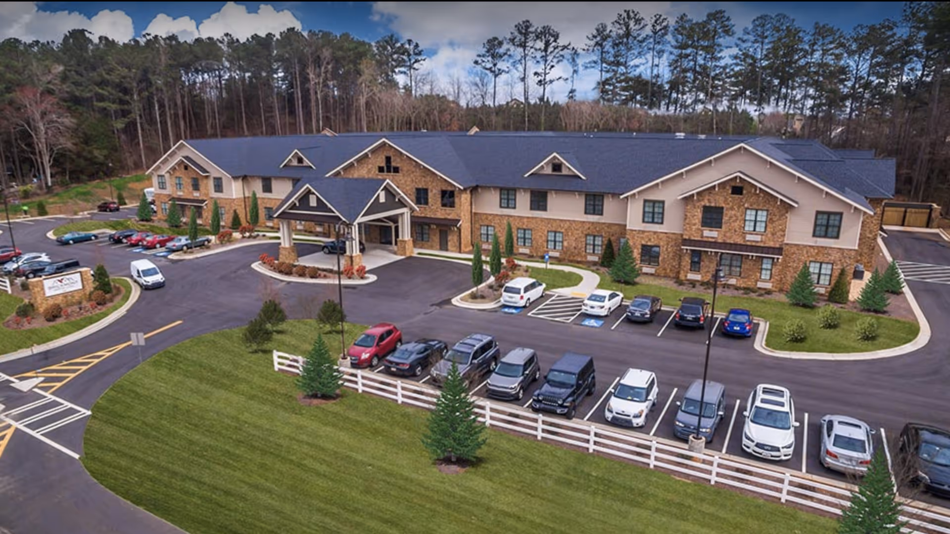 A large two-story senior living facility building with a stone and beige exterior, surrounded by a parking lot with several cars parked. The building has a covered entrance and is set against a backdrop of tall trees under a partly cloudy sky. There is a well-maintained lawn with small trees and a white fence in the foreground.