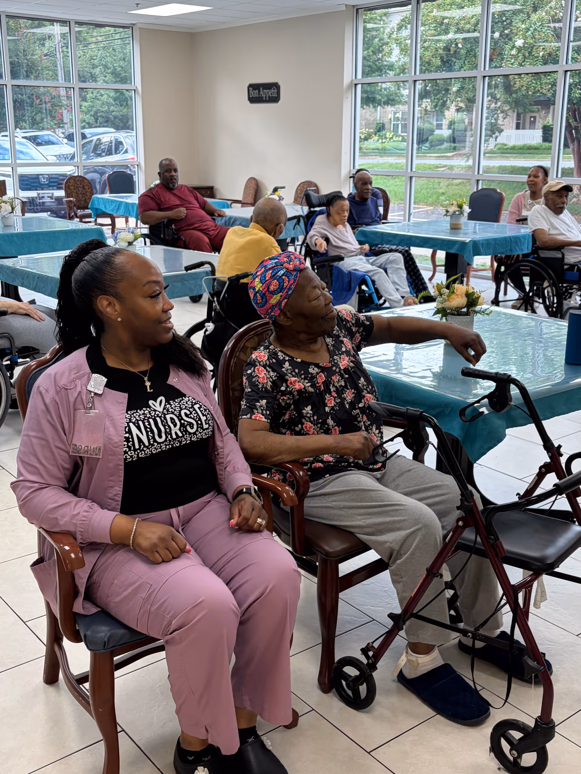 Residents sit in a bright dining area with walkers and wheelchairs.