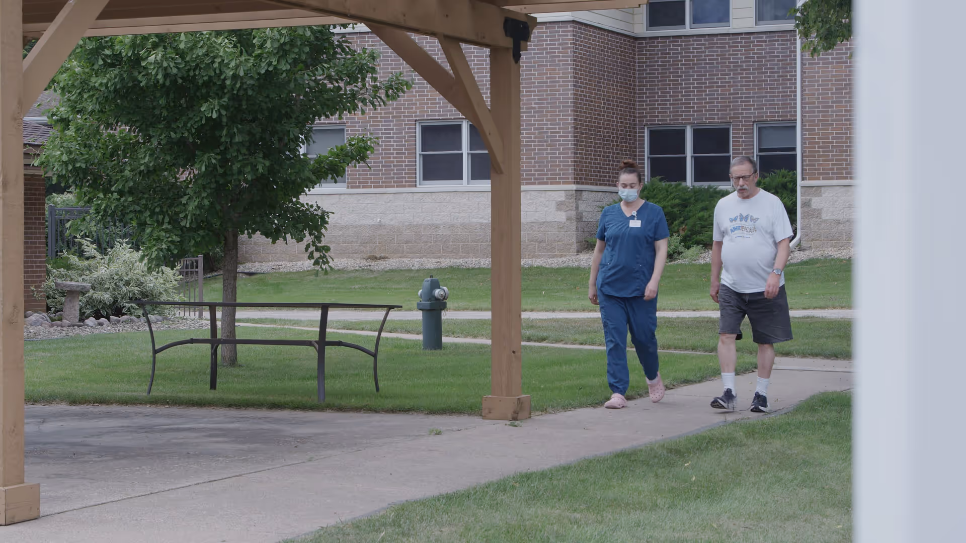 A healthcare worker wearing blue scrubs and a face mask walks alongside an elderly man in casual clothes on a paved pathway outside a brick building with green grass and trees around.