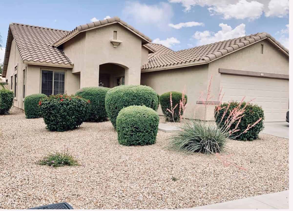 Single-story stucco house with a tiled roof, attached garage and gravel front yard with trimmed shrubs.
